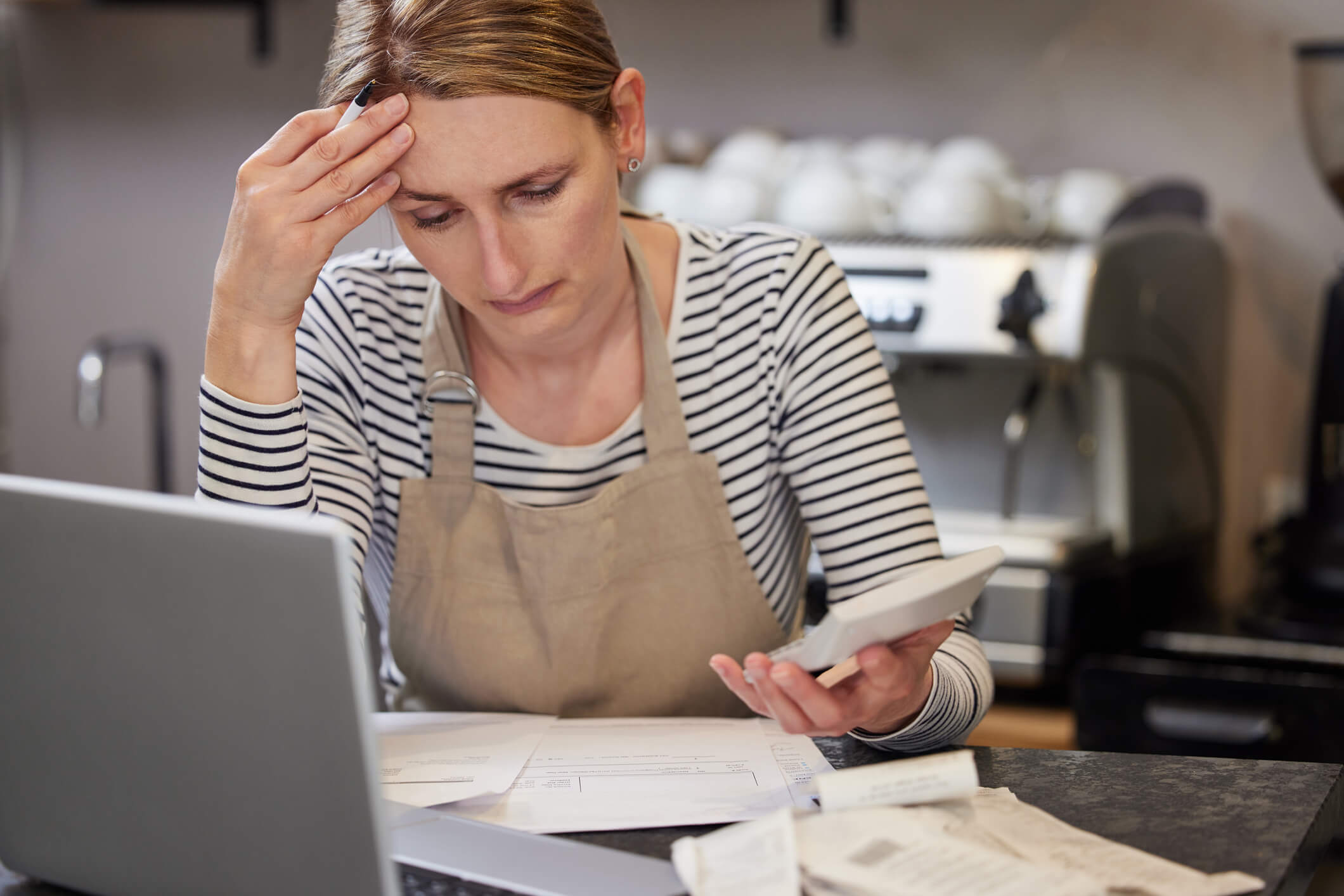 worried owner of coffee shop looking through bills using laptop and calculator